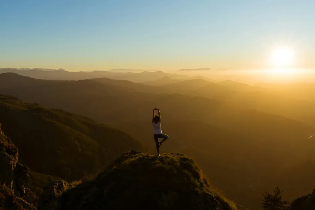 Persona che pratica una posa yoga su una montagna al tramonto, con il titolo “Gli stili di yoga – guida completa” in sovrimpressione.